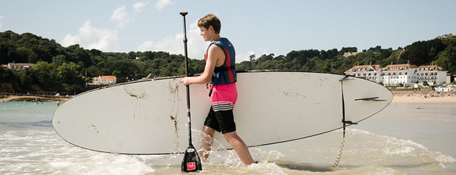 Meer und Wassersport bei einer Sprachreise für Schüler auf Jersey Ein Junge trägt ein Paddelboard ins Meer bei einer LISA! Schülersprachreise in Jersey